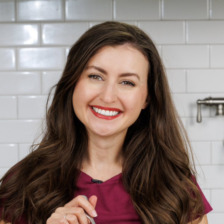 A woman with long brown hair and a bright smile stands against a background of white subway tiles. She is wearing a maroon top and appears to be in a kitchen.